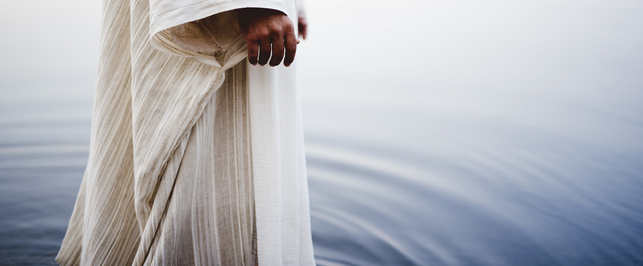 Biblical scene - of Jesus Christ walking in the water with a blurred background | E.L. Redwine Person wearing a cream striped robe standing near calm water, hand resting at the side (no face shown).